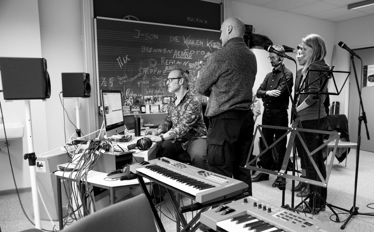 6 Personen stehen in einem Raum der Musikschule Potsdam und blicken auf einen Bildschirm, im Vordergrung sind zwei Keyboards zu sehen, im Hintergrund ein Tisch mit Musikproduktions- und Aufnahmequipment und eine beschriebene Tafel, Im Raum stehen Mirkophone und Boxen