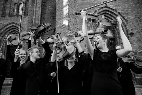 Mitglieder der Jungen Philharmonie Brandenburg halten ihre Instrumente vor einer Kirche hoch, das Foto entstand nach dem Eröffnungskonzert von Musikschulen öffnen Kirchen in der Klosterkirche Guben im April 2022
