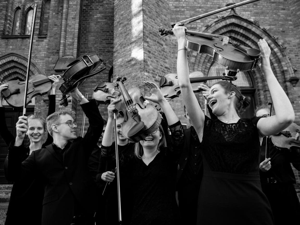 Mitglieder der Jungen Philharmonie Brandenburg halten ihre Instrumente vor einer Kirche hoch, das Foto entstand nach dem Eröffnungskonzert von Musikschulen öffnen Kirchen in der Klosterkirche Guben im April 2022